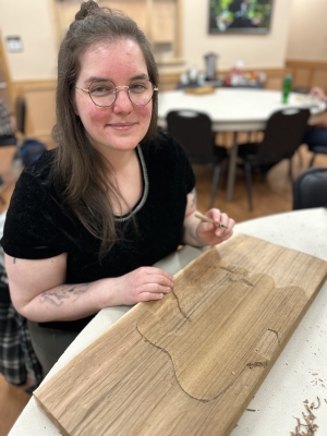Woman in black t-shirt carves wooden plank at a table