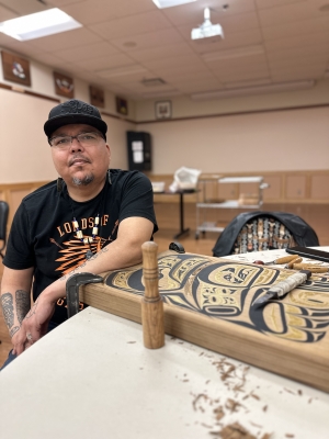 man in black t-shirt and black hat sits at a table with a wooden carving.
