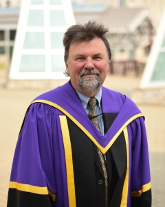 Man in academic purple, gold and black academic regalia stands outside