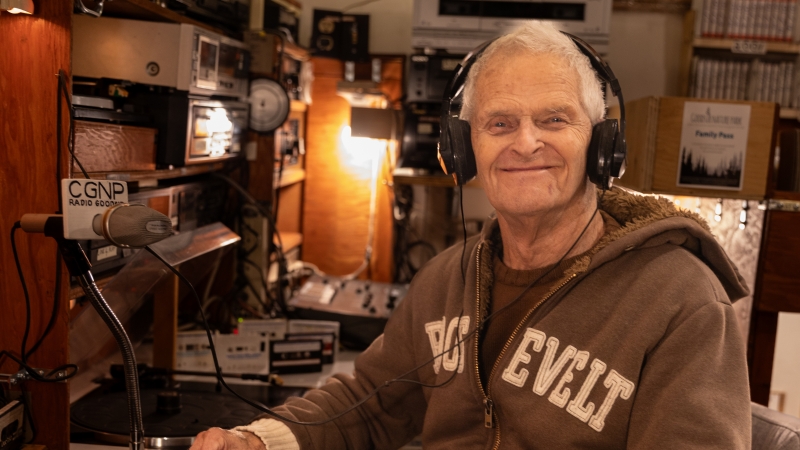Person wearing a brown hoodie and headphones sits in a radio broadcast studio with small table lamp in background.