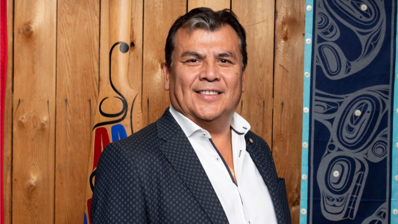 Photo of Warner Adam with wood paneling behind him and Indigenous art on the right hand side of the frame 