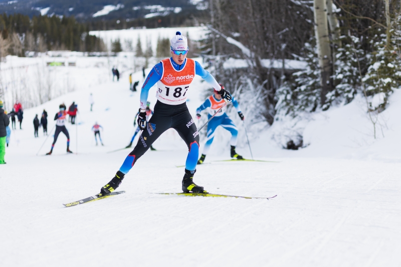 cross-country skiing athlete wearing white, light blue and navy blue race suit, skiing uphill