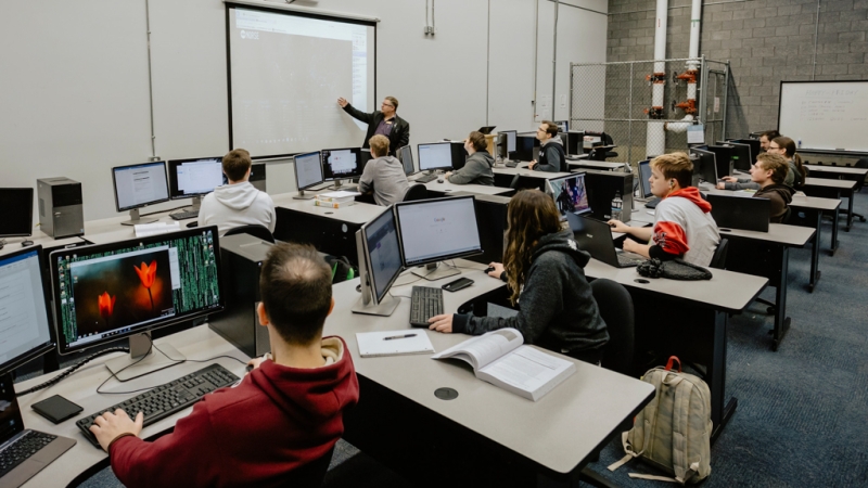 Photo shows two rows of students sitting at computer desks looking to front of room with an instructor at a projector screen.