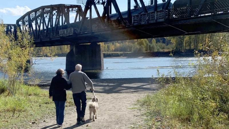 A couple of older adults walk their dog along a trail near the Fraser River. 