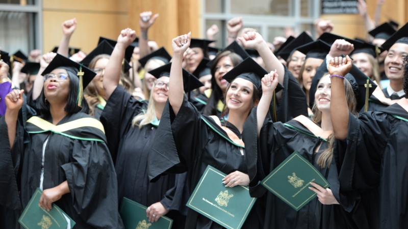 Photo shows students in academic regalia, holding diplomas and cheering.