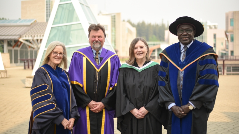Exterior photo showing four people in academic regalia with steel and glass architecture in background.