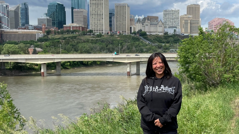 Woman wearing black hoodie with white lettering poses for photo with cityscape in the background.
