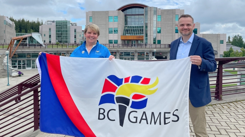 2022 BC Summer Games Host Society President Renee McCloskey and UNBC President Dr. Geoff Payne with a BC Games flag outside at UNBC's Prince George campus, library in background