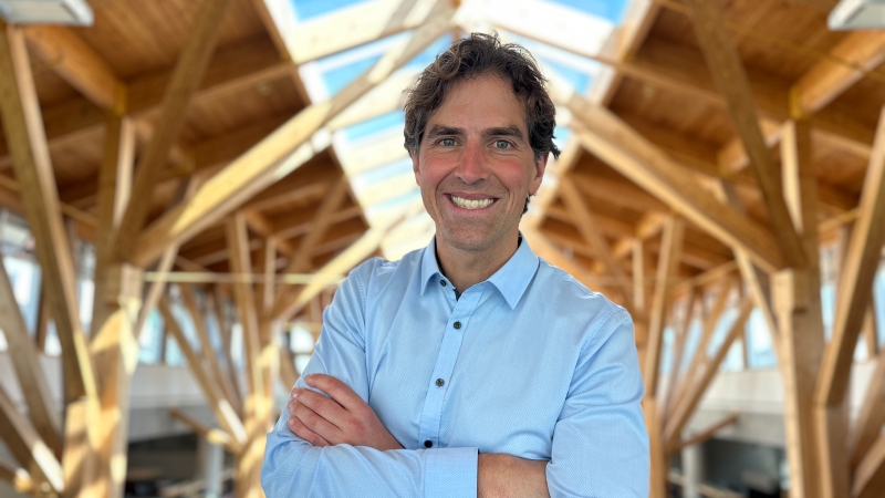 Man wearing blue dress shirt crosses arms and smiles for camera with wood beams and glass architecture in background.