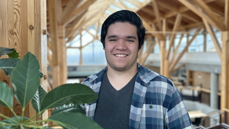 Photo shows student wearing black shirt and plaid top standing in atrium with wood beams and a plant.