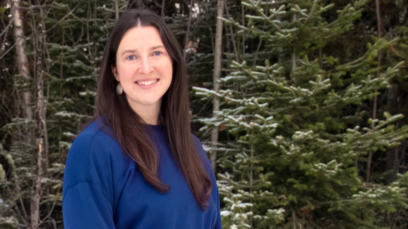 Person with long brown hair and royal blue top stands in front of coniferous trees.