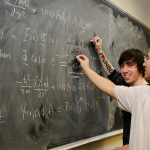 Students fill a chalkboard with physics equations.