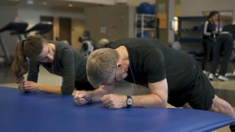 Two people, one instructor and one person living with dementia, do forearm pushups in a gym.