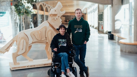 Two people in a hallway with a cut out wood Timberwolf in the background. The person on the left is in a wheelchair. 