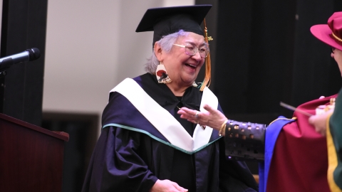 Person wearing academic mortar board and gown smiles at another person slightly off-camera who holds arms open for a congratulatory hug.