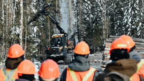 Row of people wearing orange safety vests and hard hats in foreground. In background, a yellow and black harvester-forwarder machine works in the trees.