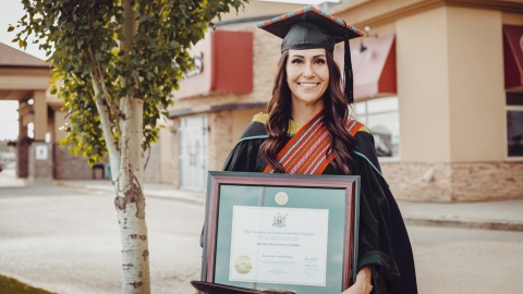 Student in black academic regalia - cap and gown - holds framed diploma while posing for photo. Tree to left side and building in background.