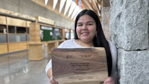 student holding wooden award plaque, leaning against rock wall