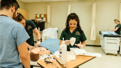 Three nursing students work on a simulation model, with one other student in the background. 